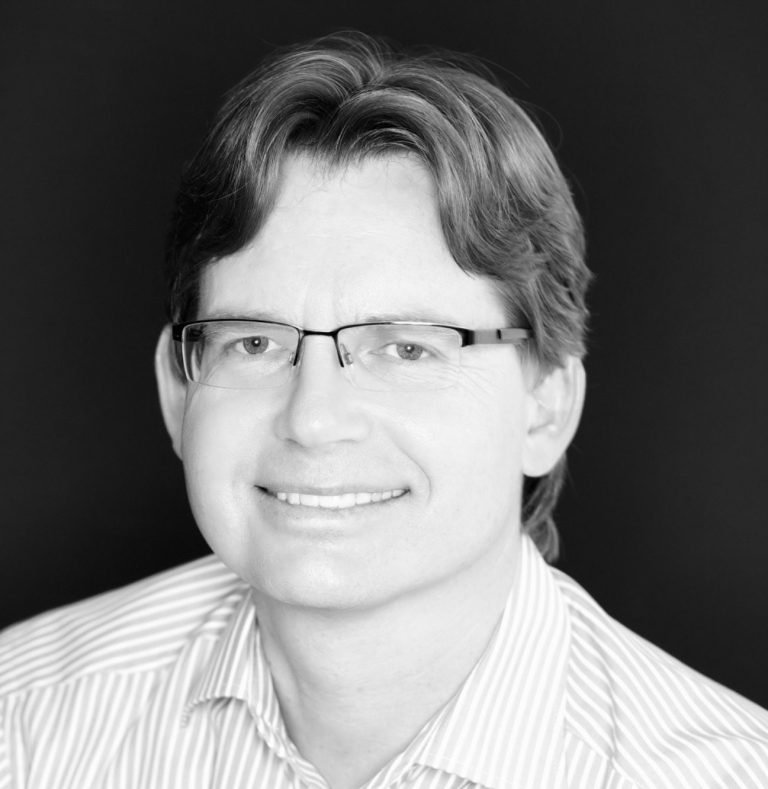 Karl Kogler – Ihr Ansprechpartner bei Fionitas-Pflege Schwarz-Weiß-Porträt von Karl Kogler, lächelnd, mit Brille und gestreiftem Hemd vor dunklem Hintergrund.